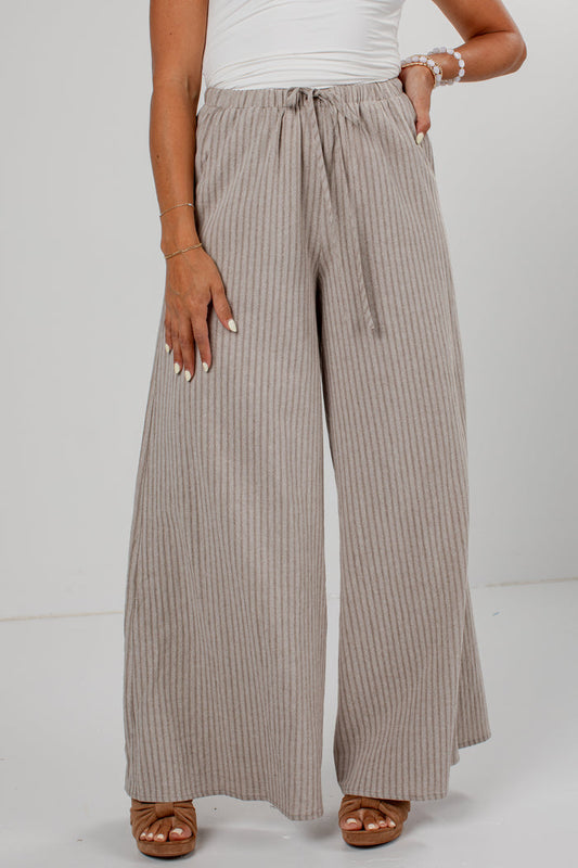A person wearing wide-leg pleated pants in a taupe-beige color with thin vertical stripes, paired with a white top and brown sandals, shown from the waist down against a light gray studio background.