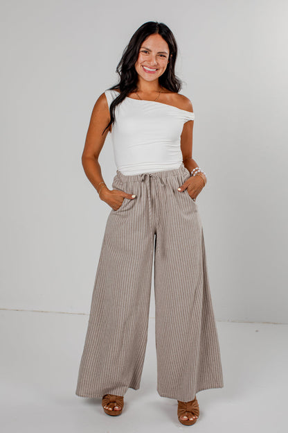 A woman modeling a white off-the-shoulder top and high-waisted, wide-leg taupe pinstripe pants against a light gray studio background.