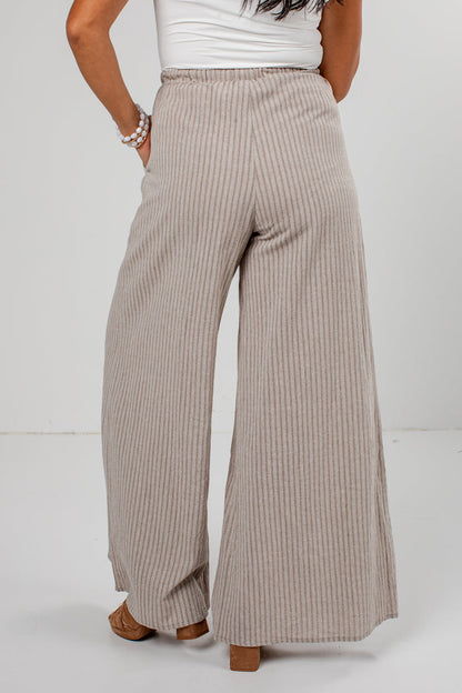 A person shown from the back wearing high-waisted, wide-leg pleated pants in a light beige and white stripe pattern, paired with a white top and brown block-heel sandals.