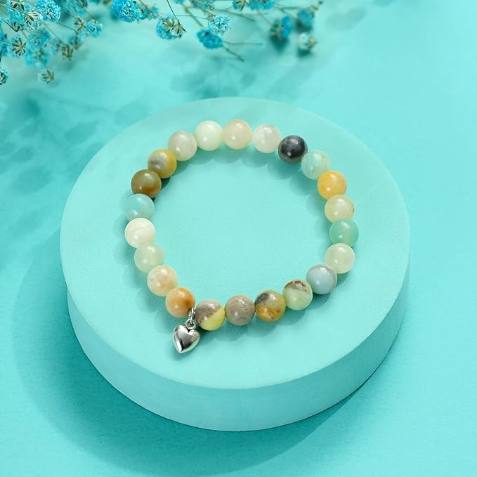 A pastel-toned gemstone beaded bracelet with a small silver heart charm placed on a turquoise round display stand with blue dried flowers in the background.