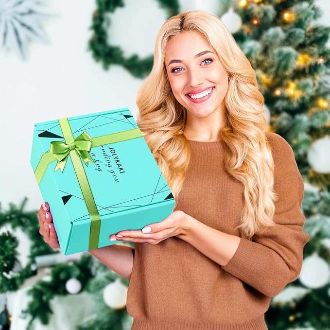 A smiling woman with long blonde hair holding a turquoise gift box with a green ribbon, standing in front of a decorated Christmas tree.