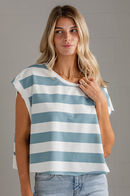 A woman wearing a loose, short-sleeve top with horizontal light blue and white stripes, paired with light-wash jeans, standing against a light gray wall.