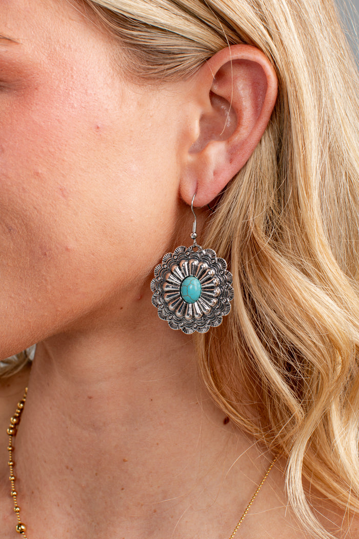 A close-up of a woman's ear wearing a silver-toned Southwestern-style dangle earring with a turquoise stone in the center.