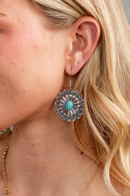A close-up of a woman's ear wearing a silver-toned Southwestern-style dangle earring with a turquoise stone in the center.