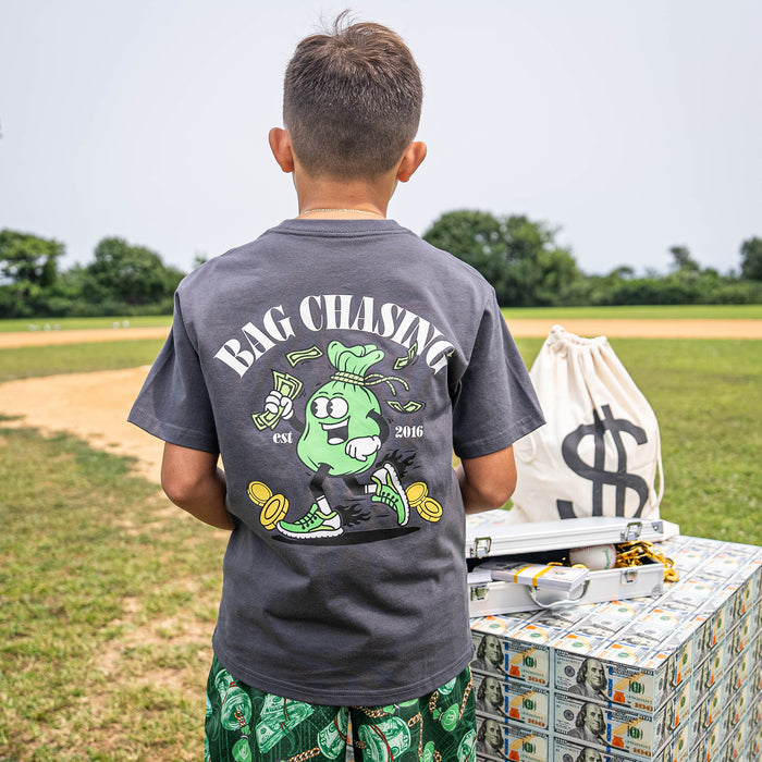 Child wearing a t-shirt with 'Bag Chasing' text and bag holding money graphic, standing near a pile of money and bags with dollar signs.