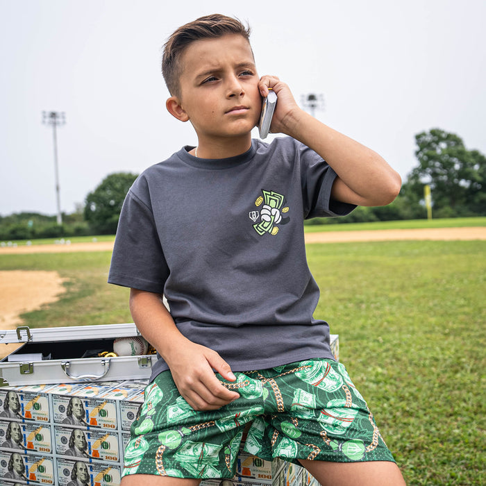 Child sitting on a pile of money with a suitcase, wearing a gray t-shirt and green money patterned shorts, outdoors.