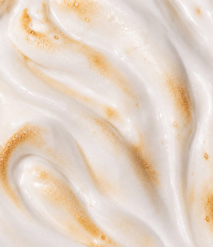 A close-up texture shot of whipped cream or whipped body butter, showing soft white peaks swirled with fine golden-brown sugar crystals.