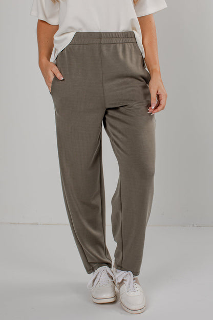 A person wearing loose, straight-leg jogger pants in a muted taupe-gray color, paired with a white t-shirt and white sneakers, standing against a light gray studio background.