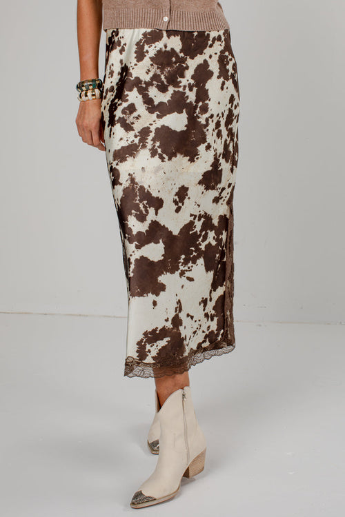 A model is shown from the waist down wearing a brown and white cow-print midi skirt with a lace hem, paired with cream-colored pointed-toe ankle boots.