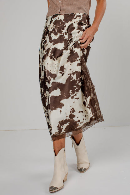 A model is shown from the waist down wearing a brown and white cow-print midi skirt with a lace hem, paired with cream-colored pointed-toe ankle boots.
