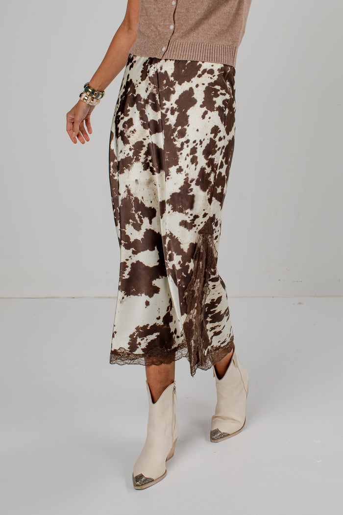 A model is shown from the waist down wearing a brown and cream cow-print skirt with a lace hem, paired with beige ankle boots and a taupe cardigan, against a plain light gray studio background.